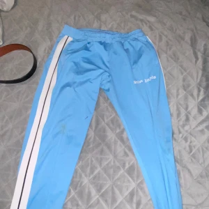 Blå mjukisbyxor med vita ränder - Säljer ett Palm angels par blå mjukisbyxor med vita ränder längs sidorna. De har en elastisk midja och ser riktigt bekväma ut för chill dagar hemma eller lätt träning. Märket är inte helt tydligt, men det står något på sidan av byxorna.