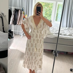 Vit långklänning från Namaste India - Säljer en superfin vit långklänning från Namaste India. Den har ett vackert paisleymönster i beige och guld, och är gjord i ett lätt och luftigt material. Klänningen har en v-ringad design med snörning framtill och volangdetaljer vid ärmslut och nederkant. Storlek L 