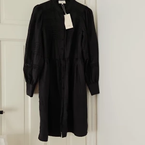 Svart klänning från Lindex - Säljer en elegant svart klänning från Levate. Oanvänd. Den har långa ärmar med vackra spetsdetaljer och knappar framtill. Perfekt för en festlig kväll eller en stilren vardag. Klänningen är i nyskick och redo att bäras med stil!