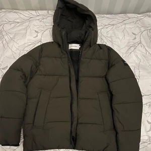 Grön pufferjacka från Calvin Klein - Säljer en snygg och varm grön pufferjacka från Calvin Klein. Jackan har en dragkedja framtill och en mysig huva för extra skydd mot kylan. Har knappt använt den.
