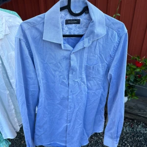 Blå skjorta från Dressmann - Säljer en snygg blå skjorta från Dressmann. Den har en klassisk krage och långa ärmar. Skjortan har ett diskret mönster på insidan av kragen. Perfekt för både vardag och fest!