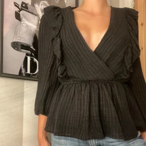 Svart omlottopp med volangdetaljer - Säljer en superfin svart omlottopp med volangdetaljer. Toppen har trekvartsärmar och en v-ringad design som ger en elegant look. Perfekt för både vardag och fest!