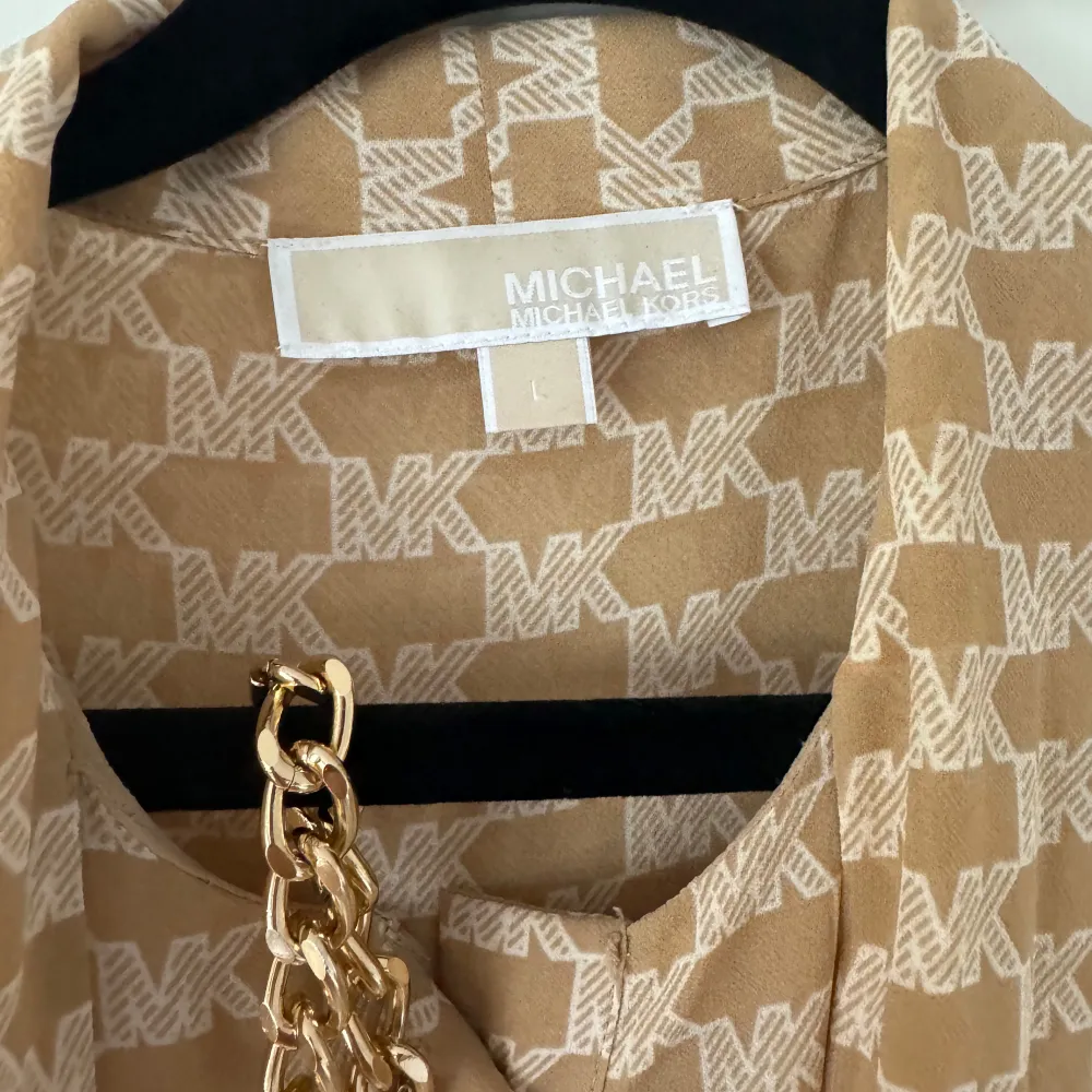 Säljer en snygg beige klänning från Michael Kors med ett coolt MK-mönster över hela tyget. Klänningen har långa ärmar och en elegant kedjedetalj som ger den en lyxig touch. Perfekt för både vardag och fest!. Klänningar.