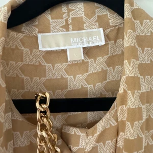 Beige klänning från Michael Kors - Säljer en snygg beige klänning från Michael Kors med ett coolt MK-mönster över hela tyget. Klänningen har långa ärmar och en elegant kedjedetalj som ger den en lyxig touch. Perfekt för både vardag och fest!