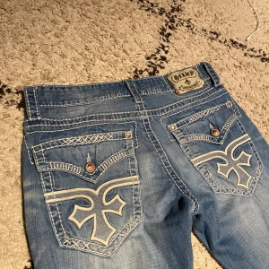 Olimp/affliction jeans - Snygga blå jeans med unika broderade detaljer på bakfickorna. De har en klassisk femficksdesign och en cool tvättad look. Perfekta för dig som vill ha något extra i garderoben.                                                       Mått ytterbenslängd 108cm, innerbenslängd 76cm och midja 47cm