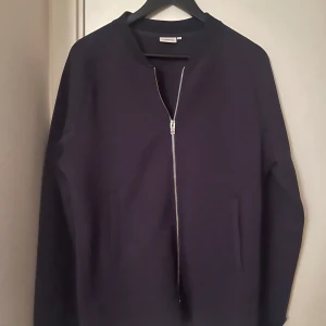 Mörkblå zip från J.Lindeberg - Snygg mörkblå zip från J.Lindeberg med dragkedja framtill. Zipen har en stilren design med långa ärmar och är perfekt för en cool look. Passar bra till både jeans och chinos. Hör av dig vid fråga.
