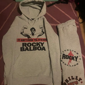 Grå Rocky hoodie  - Säljer en grå Rocky tracksuit. Perfekt för fans av den ikoniska filmen. Hoodien har en klassisk passform med känguruficka och ribbade muddar.