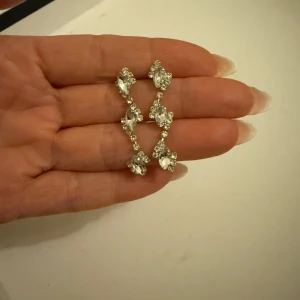 Glittriga örhängen i silver - Eleganta och glittriga örhängen i silver med en böljande design. Perfekta för att ge en extra touch av glamour till din outfit. De har en rad av gnistrande stenar som fångar ljuset vackert.