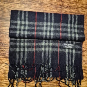 Rutig halsduk från Burberry - Säljer en elegant rutig halsduk från Burberry i 100% kashmir. Den har en klassisk design med mörk blåa, grå och röda rutor samt fransar i ändarna. Perfekt för att hålla dig varm och stilren under kyliga dagar.