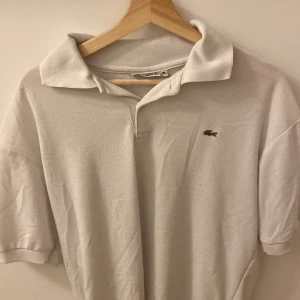 Vit pikétröja från Lacoste - Säljer en klassisk vit pikétröja från Lacoste med den ikoniska krokodilloggan på bröstet. Tröjan har korta ärmar och en stilren krage. Perfekt för en avslappnad och stilren look. L/XL