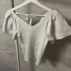 Vit ribbad topp med puffärmar - Säljer en stilren vit ribbad topp med v-ringning och puffiga kortärmar. Toppen har en knytning i ryggen som ger en extra detalj. Perfekt för en trendig look!