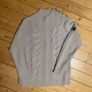 Grå stickad tröja från Balmain - Säljer en stilren grå stickad tröja från Balmain Yacht Edition. Tröjan har ett snyggt flätmönster och knappar på axeln. Perfekt för kyliga dagar med sin höga krage och långa ärmar. Ett måste för garderoben! 🧶