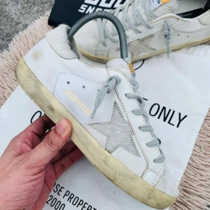 Golden Goose vita sneakers - Säljer ett par vita Golden Goose sneakers med grå snörning och en ikonisk stjärndetalj på sidan. Skorna har en vintage-look med slitna detaljer och en gulnad sula för en unik stil. Perfekta för dig som gillar en avslappnad och trendig look.