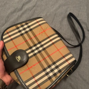 Rutig axelväska från Burberry - Säljer en klassisk rutig axelväska från Burberry i beige, svart och röd. Väskan har en justerbar axelrem och en stilren design med läderdetaljer. Perfekt för att ge din outfit en touch av lyx.
