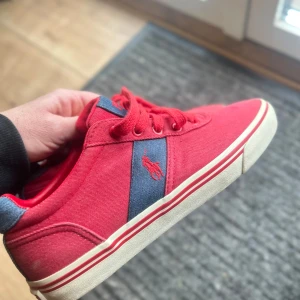 Röda sneakers från Polo Ralph Lauren - Snygga röda sneakers från Polo Ralph Lauren med blå detaljer och vit sula. Skorna har snörning och en klassisk design med märkets logga på sidan. Perfekta för en avslappnad stil.