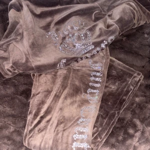 Brun velour hoodie och byxor från Juicy Couture - Säljer en brun velour hoodie och matchande byxor från Juicy Couture. Hoodien har en glittrig Juicy Couture-logga med en krona på baksida. Byxorna har en liknande glittrig logga längs benet. Perfekt för en avslappnad och stilren look. Knappt använda