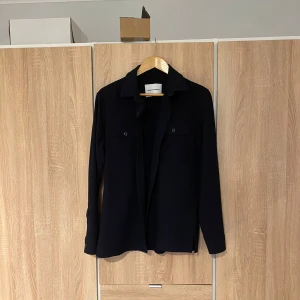 Mörkblå overshirt från shaping new tomorrow - Snygg mörkblå overshirt från shaping new tomorrow med klassisk krage och knappar framtill. Perfekt för lager-på-lager-stil. Två bröstfickor med lock ger en stilren look. Passar både till vardags och mer uppklädda tillfällen.