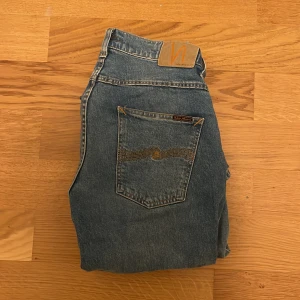 Blå jeans från Nudie Jeans - Snygga blå jeans från Nudie Jeans med klassisk femficksdesign och en cool broderad detalj på bakfickorna. Perfekta för en avslappnad stil med en modern touch. Midjan är normal och passformen är straight. Varan har används 2 gånger.
