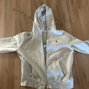 grå vit hoodie från Ralph Lauren - Säljer en stilren grå vit hoodie från Ralph Lauren med dragkedja och huva. Tröjan har långa ärmar och en liten broderad logga på bröstet. Perfekt för en avslappnad look.