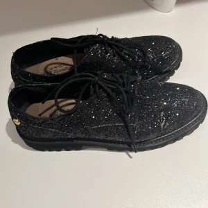 Svarta glittriga skor med snörning - Snygga svarta skor med glittrig yta och snörning. Perfekta för att ge en extra touch till din outfit. Skorna har en låg sula och är dekorerade med små glittrande detaljer.