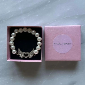 Pärlarmband med hjärtformad detalj  - Elegant pärlarmband med en glittrig hjärtformad detalj i mitten. Armbandet består av vita pärlor och har en vacker design som passar perfekt för speciella tillfällen.