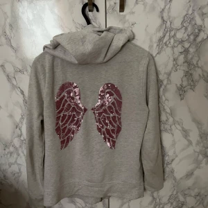 Victoria’s Secret Angel Hoodie - Säljer en snygg grå hoodie från Victoria's Secret med rosa paljettvingar på ryggen. Hoodien har dragkedja med en liten vinge som detalj. Perfekt för en avslappnad stil med en touch av glamour.