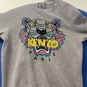 Grå sweatshirt från Kenzo - Säljer en grå sweatshirt från Kenzo med ett stort, färgglatt tigerbroderi på framsidan. Tröjan har rund hals och är kortärmad. Perfekt för en cool och avslappnad stil.