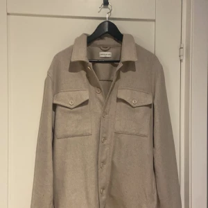 Beige overshirt från These Glory Days - Säljer en stilren beige overshirt från These Glory Days i storlek medium. Den har knappar framtill och två bröstfickor med lock. Perfekt för lager-på-lager-stil och passar till många olika outfits. Ny pris runt 2000 kr 