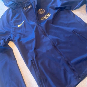 Blå jacka från Nike med PSG-logga - Säljer en blå Nike-jacka med PSG-logga och Qatar Airways-tryck. Jackan har dragkedja och långa ärmar, perfekt för fotbollsfans. Den är tillverkad i Dri-FIT-material för optimal komfort.