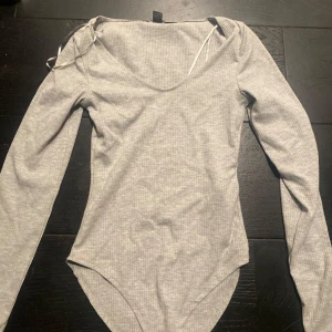 Grå ribbad bodysuit - Säljer en stilren grå ribbad bodysuit med långa ärmar och rund hals. Perfekt för lager-på-lager eller att bära som den är. Den har en bekväm passform och är gjord i ett mjukt material.