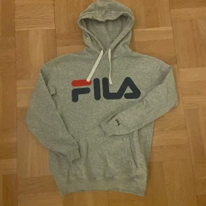 Grå hoodie från FILA - Säljer en grå hoodie från FILA med stor logga på bröstet. Tröjan har en klassisk känguruficka och justerbar huva med snören. Perfekt för en avslappnad stil.