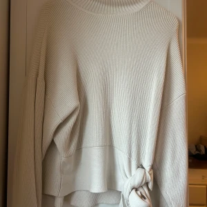 Vit ribbad polotröja från Zara - Säljer en stilren vit ribbad polotröja från Zara. Tröjan har en knytning i sidan som ger en unik touch. Perfekt för kyligare dagar med sin höga krage och långa ärmar. Ett måste i garderoben för en chic look!