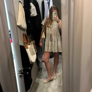 Blommig kortklänning - Säljer en söt och luftig kortklänning med blommigt mönster. Klänningen har korta ärmar och en dekorativ tofs framtill. Perfekt för en avslappnad och stilren look.