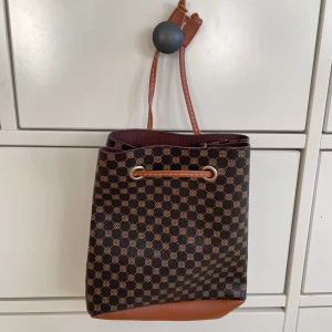 Mönstrad axelväska med bruna detaljer - Snygg axelväska med ett klassiskt mönster i brunt och svart. Väskan har en justerbar axelrem och en praktisk dragsko för enkel öppning och stängning. Perfekt för att bära dina dagliga nödvändigheter med stil.