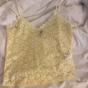 Gul spetslinne - Säljer ett gulligt gult spetslinne med smala axelband. Linnet har ett blommigt mönster och en v-ringad design. Perfekt för varma dagar eller som ett lager under en annan topp.