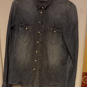 Blå jeansskjorta från Denim by Lindex - Snygg blå jeansskjorta från Denim by Lindex i storlek 36. Skjortan har långa ärmar och knappar framtill samt två bröstfickor med knappstängning. Perfekt för en avslappnad stil.