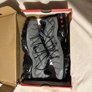 Grå Nike TN - Snygga grå Nike sneakers med svart sula och synliga luftkuddar i hälen. Skorna har ett stickat mönster och snörning framtill. Perfekta för en sportig look samt perfekta i alla väder.