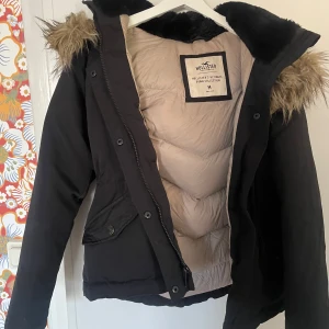 Svart dunjacka från Hollister - Säljer en svart dunjacka från Hollister med fuskpäls på huvan. Jackan har en dragkedja och knappar framtill samt två fickor med knappar. Insidan är ljus och quiltad för extra värme. Perfekt för kyliga dagar!