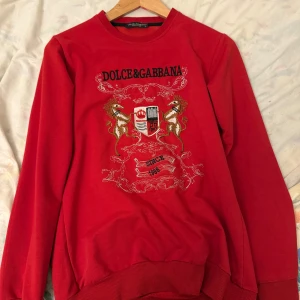 Röd tröja från Dolce & Gabbana - Säljer en snygg röd tröja från Dolce & Gabbana med ett broderat emblem på framsidan. Tröjan har långa ärmar och ribbade muddar vid ärmslut och nederkant. Perfekt för dig som vill ha något stilrent och unikt. Hör av dig vid intresse!