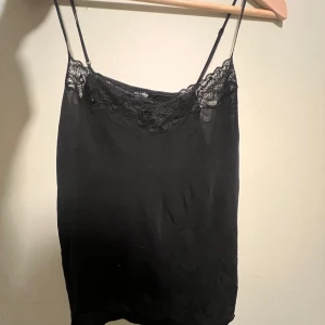 Svart linne från Change Lingerie - Säljer ett elegant svart linne från Change Lingerie med spetsdetaljer vid halsringningen. Linnet har tunna axelband och en mjuk, följsam passform. Perfekt för en stilren look.