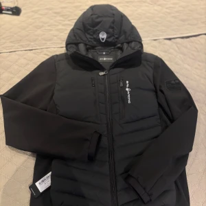 Svart jacka från Sail Racing - Säljer en snygg svart jacka från Sail Racing med huva och dragkedja. Jackan har flera praktiska fickor med dragkedjor och en stilren design. Perfekt för kyligare dagar.