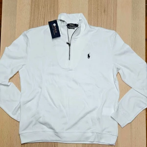 Vit tröja med dragkedja från Polo Ralph Lauren - Säljer en stilren vit tröja från Polo Ralph Lauren med halv dragkedja och klassisk logga på bröstet. Perfekt för en avslappnad men ändå snygg look. Tröjan har långa ärmar och är tillverkad i ett mjukt material.