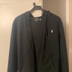 Svart hoodie från Ralph Lauren - Säljer en stilren svart hoodie från Ralph Lauren med dragkedja och justerbar huva. Den har en broderad logotyp på bröstet och praktiska fickor framtill. Perfekt för en avslappnad look.