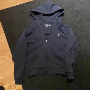 Mörkblå hoodie från Ralph Lauren - Säljer en stilren mörkblå hoodie från Ralph Lauren med dragkedja och det klassiska broderade logotypen på bröstet. Perfekt för en avslappnad stil. 🐎 Den är i helt nytt skick