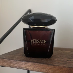 Versace Crystal Noir Eau de Parfym 30ml, hälften kvar - Elegant parfymflaska från Versace i en mörk, nästan svart nyans. Flaskan har en unik, facetterad kork som ger en lyxig känsla. Perfekt för den som vill ha en sofistikerad doftupplevelse.