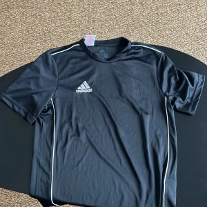 Svart fotbollströja från Adidas - Snygg svart fotbollströja från Adidas med korta ärmar och vita detaljer längs sidorna. Perfekt för träning eller match. Tillverkad i lätt och andningsbart material för optimal komfort.