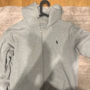 Grå hoodie från Ralph Lauren - Säljer en stilren grå hoodie från Ralph Lauren med dragkedja och det klassiska logotypbroderiet på bröstet. Perfekt för en avslappnad look. Den har långa ärmar och en bekväm passform.