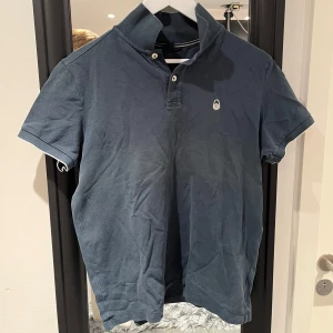 Blå pikétröja från sail racing - Snygg blå pikétröja med korta ärmar och klassisk krage. Tröjan har en liten logotyp på bröstet och knappar vid halsen. Perfekt för en avslappnad stil.