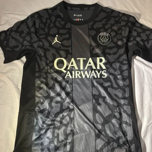 Svart och grå PSG fotbollströja - Snygg PSG fotbollströja i svart och grått med kamouflagemönster. Tröjan har korta ärmar och det ikoniska Jordan-logotypen på bröstet. Perfekt inför sommaren 