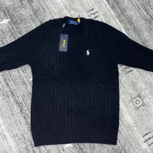 Svart stickad tröja från Polo Ralph Lauren - Säljer en klassisk svart stickad tröja från Polo Ralph Lauren med det ikoniska vita logotypbroderiet på bröstet. Tröjan har en rund halsringning och långa ärmar, perfekt för kyligare dagar. Storlek M men det funkar jätte bra som storlek S